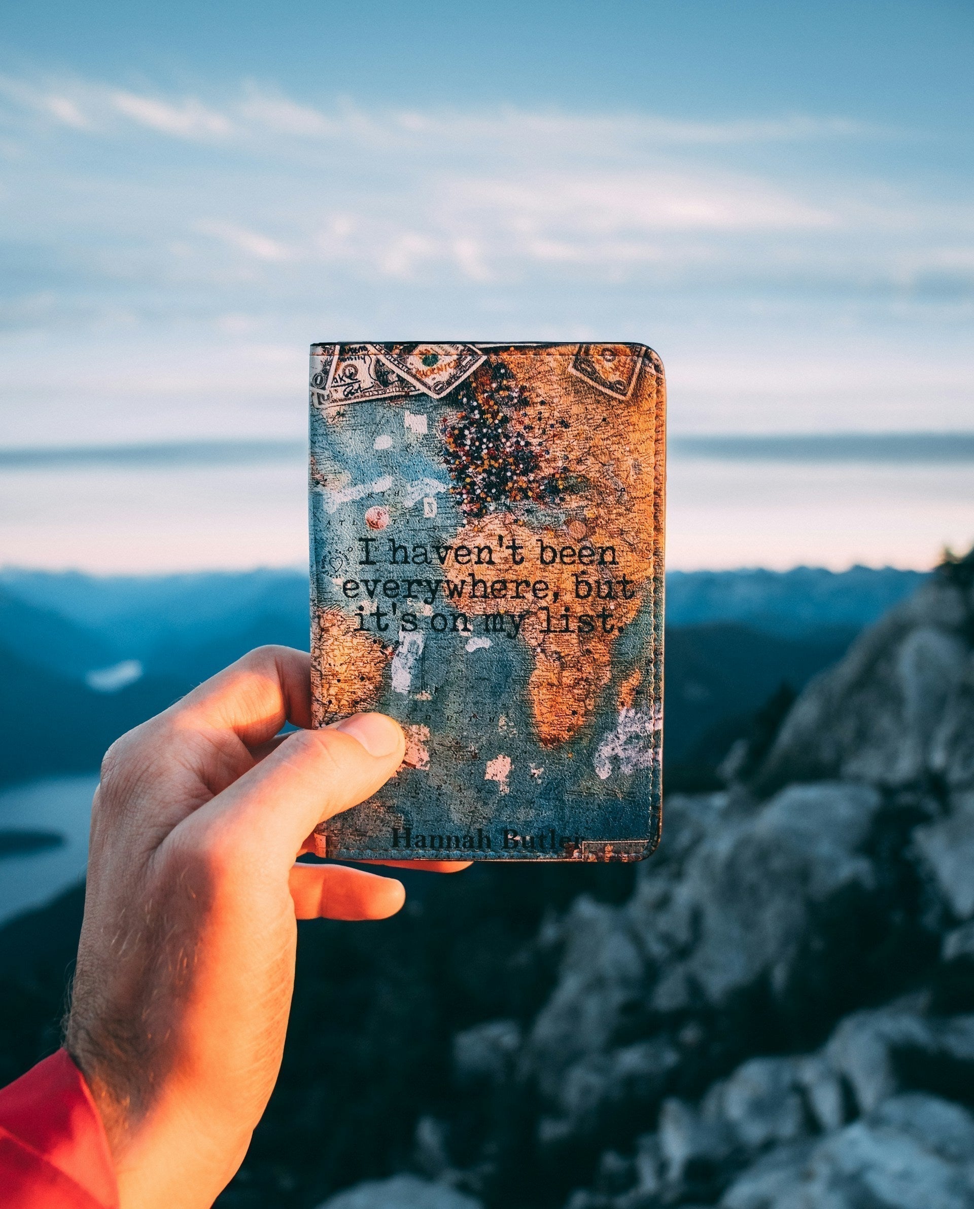 Étui de voyage avec la citation "I haven't been everywhere, but it's on my list", tenu devant un paysage de montagnes et de lac