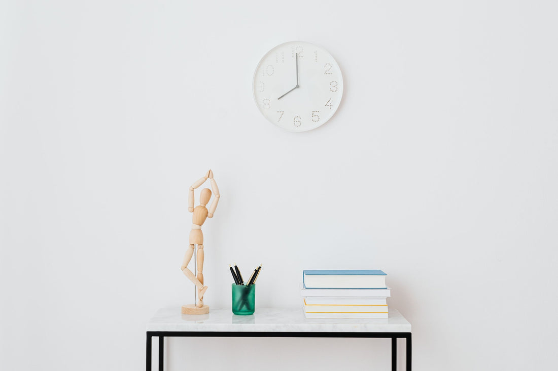 Horloge murale blanche au-dessus d'une figurine en bois en posture de yoga, entourée de stylos et d'une pile de livres sur une table d'appoint en marbre.