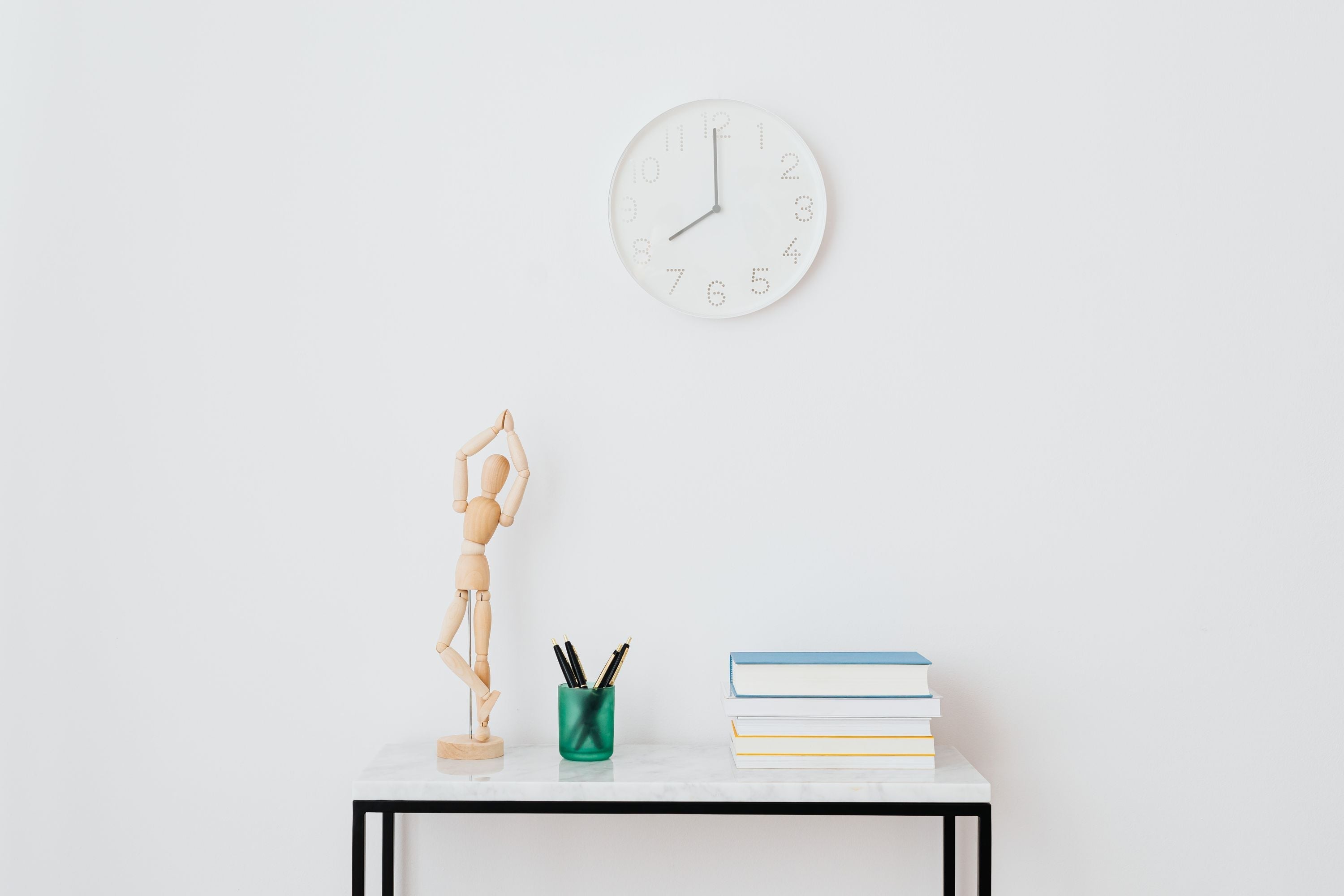 Horloge murale blanche au-dessus d'une figurine en bois en posture de yoga, entourée de stylos et d'une pile de livres sur une table d'appoint en marbre.